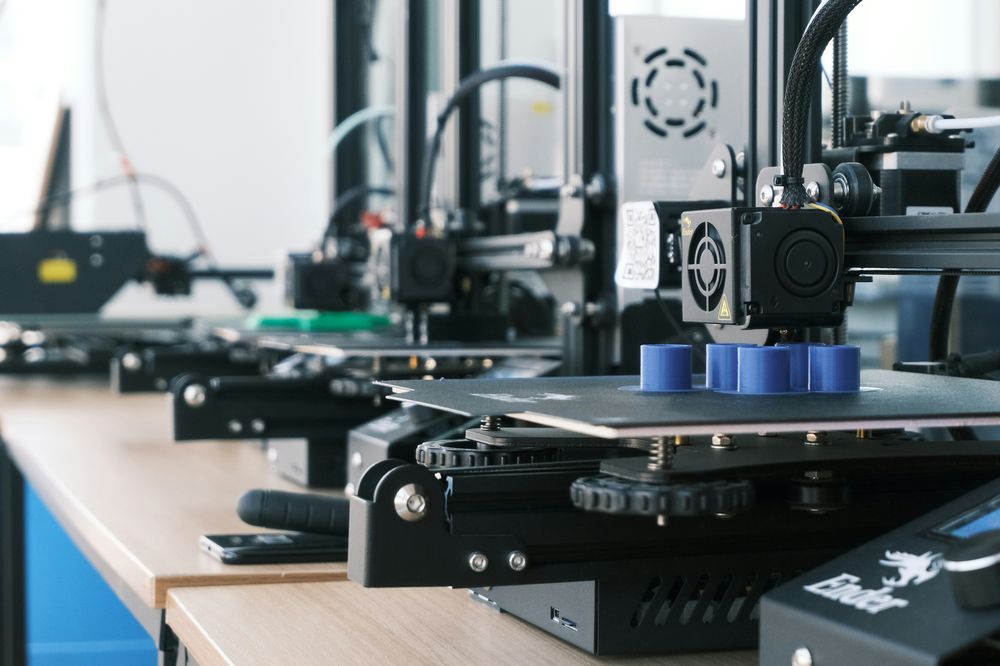 A row of 3D printers printing blue plastic cylinders on a wooden workbench.