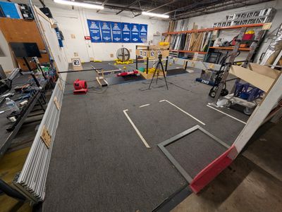 The practice field from one end, with a table of electronics and a monitor in the foreground and robots, AprilTag targets, and yellow game pieces at the far end.