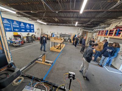 Students and mentors gather on the carpeted practice field, with wooden field element mockups and FIRST Robotics Competition award banners on the back wall.