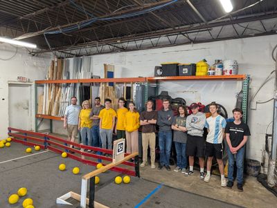 A group of students and mentors, several in matching yellow shirts, poses beside the field wall with yellow game pieces on the carpet.