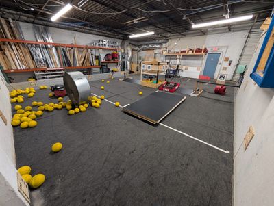 Yellow game pieces scattered across the field around a large metal drum and a tilted black platform, with a robot at the far end.