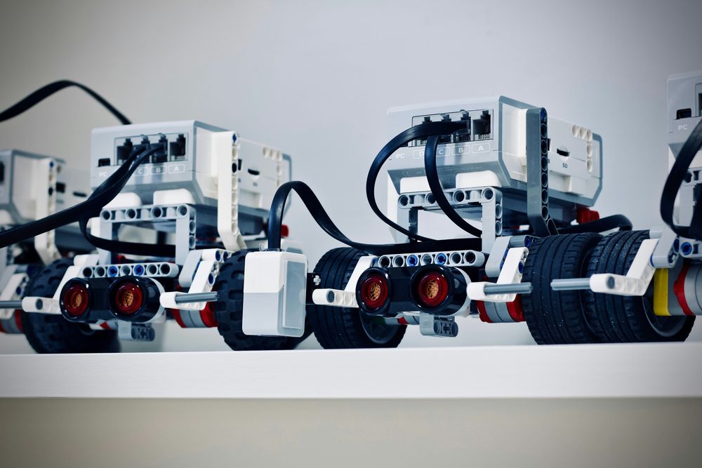 A row of LEGO robots on a white shelf, each with a programmable brick, wheels, and a distance sensor on the front.